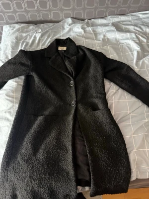 Svart bouclé kappa med knappar - Stilren svart kappa i bouclétyg med två stora knappar framtill och klassisk krage. Kappan har raka ärmar och två fickor framtill. Perfekt för dig som vill ha en tidlös och trendig look till hösten eller vintern.