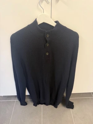 Mörkblå stickad tröja med knappar från Bruun & Stengade, Stl M/L - Snygg mörkblå stickad tröja med hög krage och tre knappar framtill. Tröjan har långa ärmar och ribbade muddar vid ärmslut och nederkant. Perfekt för kyligare dagar och enkel att matcha med olika outfits.