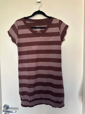 Randig brun t-shirt från Lacoste S-M - Snygg brun och ljusbrun randig t-shirt från Lacoste med v-ringning och korta ärmar. Klassisk krokodil-logga på bröstet. Tillverkad i mjuk bomull som känns skön mot huden. Perfekt för en avslappnad och stilren look.
