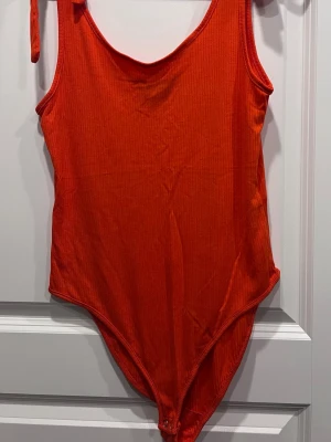 Röd/orange ribbad body från Primark - Säljer en snygg röd/orange body från Primark i ribbat tyg. Bodyn har smala knyt axelband, rundad ringning och knäppning i grenen. Perfekt att styla till jeans eller kjol för en trendig look. Materialet är mjukt och stretchigt för skön passform.