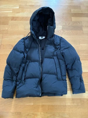 Svart pufferjacka från ARKET - Säljer en svart pufferjacka från ARKET med fyllning av 100% återvunnet dun och fjädrar (RE:DOWN). Jackan har huva med dragsko, två stora fickor framtill och dragkedja. Perfekt för kalla vinterdagar och riktigt skön att bära.