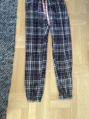 Rutiga pyjamasbyxor med  - Mysiga pyjamasbyxor i mjukt tyg med rutigt mönster i mörkblått, vitt och gråt å rosa. Byxorna har en rosa dragsko i midjan för justerbar passform och resår vid bensluten. Perfekta för chillkvällar hemma.