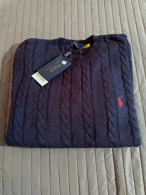 Mörkblå kabelstickad tröja Polo Ralph Lauren - Kabelstickad mörkblå tröja från Polo Ralph Lauren med rund halsringning och klassisk röd broderad logga på bröstet. Tröjan har långa ärmar och ribbade muddar. Perfekt för dig som gillar stilren och tidlös design.