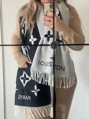 Vuitton svartvit halsduk - Snygg svartvit halsduk från Vuitton med ikoniska monogram och texten PARIS. Halsduken har långa fransar i ändarna och är tillverkad i ett mjukt material som känns lyxigt. Perfekt accessoar för att lyfta din outfit med en exklusiv touch.