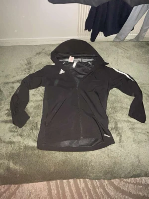 Svart hoodie med dragkedja från Adidas - Svart hoodie från Adidas med dragkedja framtill och vit rand på ena ärmen. Klassisk Adidas-logga på bröstet och huva. Tillverkad i ett lätt och sportigt material, perfekt för träning eller chill. Snygg och enkel design som funkar till det mesta. 
