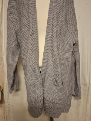 Ljusgrå stickad cardigan med fickor - Mysig ljusgrå stickad cardigan med öppen front och två stora fickor framtill. Koftan har lång ärm och ribbade muddar vid ärmslut och nederkant. Perfekt för lager på lager och chill dagar.