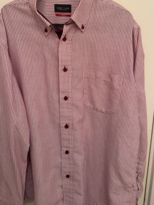 Randig skjorta från Park Lane - Säljer en klassisk skjorta från Park Lane med smala röda och vita ränder. Skjortan har lång ärm, bröstficka och röda knappar. Tillverkad i mjuk bomull och har en normal passform. Perfekt för dig som gillar stilrena detaljer.