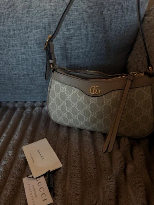 Beige axelremsväska från Gucci - Snygg beige axelremsväska från Gucci med klassiskt GG-monogram i canvas och detaljer i ljusbrunt läder. Väskan har guldfärgad logga framtill, dragkedja upptill och justerbar rem. Perfekt för dig som gillar lyxiga accessoarer med ikonisk design.