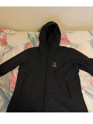 Svart vindjacka från Haglöfs med huva - Snygg svart vindjacka från Haglöfs med hel dragkedja, två fickor med dragkedja och justerbar huva. Perfekt för blåsiga dagar och outdoor-aktiviteter. Diskret logga på bröstet och stilren design.