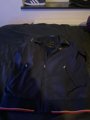 Mörkblå vindjacka från Sail Racing - Mörkblå vindjacka från Sail Racing med dragkedja framtill, två fickor med dragkedja och ribbade muddar med röd och vit rand. Jackan har en sportig look och meshfoder på insidan. Perfekt för blåsiga dagar.