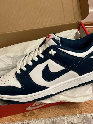 Nike Dunk - Snygga Nike Dunk Low sneakers i marinblått och vitt läder. Klassisk design med vit bas, blå overlays och vit sula. Ikonisk Swoosh-logga på sidan och röda detaljer på plösen. Perfekta för dig som gillar streetwear och sportig stil.