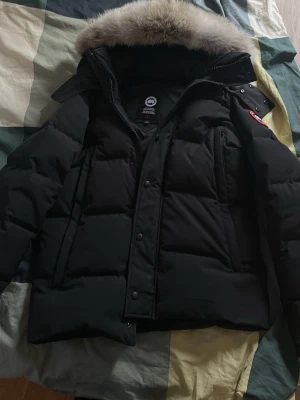 Canada goose wyndham parka äkta - Säljer en svart parkas från Canada Goose med klassisk röd och vit logotyp på ärmen. Jackan har en stor huva med fluffig pälsdetalj och dragkedja framtill. Perfekt för kalla dagar och riktigt snygg till vinterlooken.