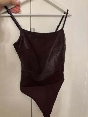 Vinröd skinmbody - Säljer en vinröd body med smala justerbara axelband och fyrkantig urringning. Materialet har en läderlik finish framtill och stretchig tyg baktill. I fejk skinn, strl xs men stretchig, endast testad 