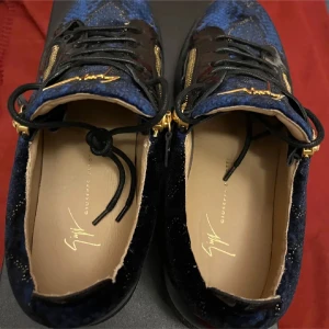 Blåa sneakers från Giuseppe Zanotti - Snygga blåa sneakers från Giuseppe Zanotti med svarta lackdetaljer, guldfärgade dragkedjor och signaturlogga på plösen. Skorna har snörning och en platt sula. Materialmix av textil och lack ger en lyxig känsla. Pris kan diskuteras vid snabb affär