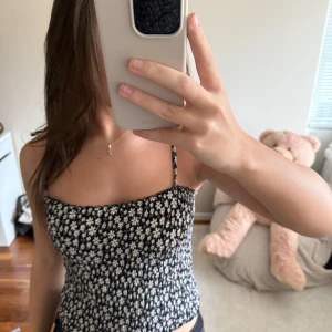 Svart blommig croppad linnetopp - Trendig svart croppad linnetopp med vita små blommor och smala axelband. Toppen har en tight passform och är perfekt för varma dagar. Materialet känns mjukt och lätt mot huden.