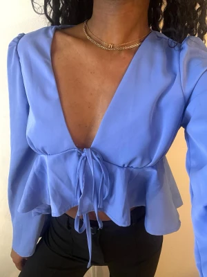 Blå volangblus med knytband - Supersnygg blå blus med djup v-ringning och volang nedtill. Blusen har långa ärmar och ett knytband i midjan som ger en feminin siluett. Tillverkad i ett lätt, flowy tyg som känns skönt mot huden. Perfekt att styla med jeans eller kjol.