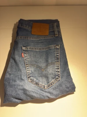 Levi's blå raka jeans - Klassiska blå Levi's jeans med rak passform och fem fickor. Jeansen har en ljus tvätt och detaljer i kopparfärg vid fickorna. Ikonisk läderpatch bak i midjan och röd Levi's-tag på bakfickan. Perfekta för en avslappnad och tidlös stil. Aldrig använda.