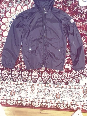 Mörklila vindjacka från Moncler - Snygg mörklila vindjacka från Moncler med huva och dragkedja framtill. Jackan har två fickor med tryckknappar och Moncler-logga på ärmen. Materialet är glansigt och lätt, perfekt för blåsiga dagar. Huva med dragsko och elastiska muddar vid ärmslut.