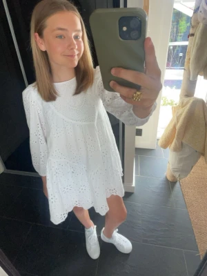 Vit kort klänning med broderade detaljer - Superfin vit kort klänning med broderade hålmönster och vida långärmar. Klänningen har en lös passform och rund halsringning, perfekt för varma dagar. Snygg och trendig stil som passar till många tillfällen.