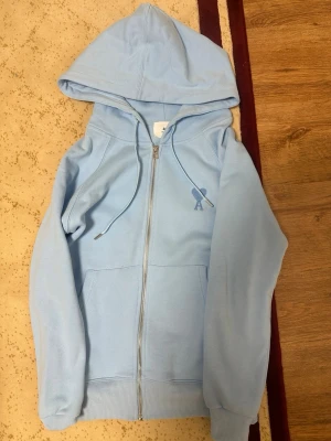 Ljusblå zip up från AMI PARIS - Snygg ljusblå hoodie från AMI Paris med dragkedja framtill, huva med dragsko och fickor. Tillverkad i mjuk bomull och har det klassiska AMI-hjärtat broderat på bröstet. Perfekt för en chill och trendig look.