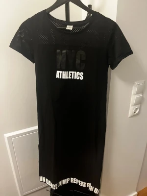 Svart nattlinne NYC Athletics - Svart nattlinne med texten 'NYC ATHLETICS' framtill och vit text längs nederkanten. Kortärmad modell med små håldetaljer upptill för extra stil. Perfekt för en avslappnad kväll hemma.