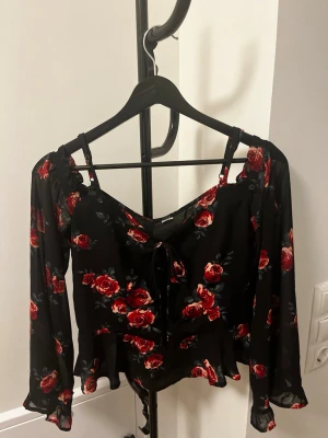 Svart blommig off shoulder blus med snörning - Supersöt svart off shoulder blus med röda rosor, långa ärmar och volangkant. Toppen har smala axelband och snörning framtill för extra detalj. Perfekt för dig som gillar romantisk stil och vill sticka ut lite extra.