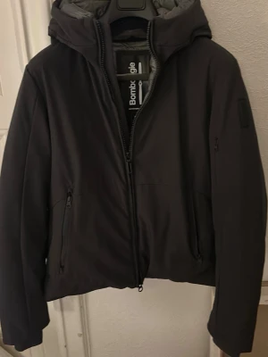 Svart jacka från Bombogie - Snygg svart jacka från Bombogie med huva och dragkedja framtill. Jackan har två sidofickor och även en ficka inuti jackan och en på armen. En diskret logotyp på ärmen. Materialet är slätt och vattenavvisande samt är det tjockt, vilket gör det perfekt för blåsiga och kalla dagar. Pris kan diskuteras!