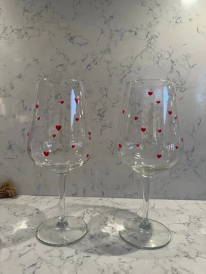Två vinglas med röda hjärtan - Säljer två stilrena vinglas i klart glas, dekorerade med små röda hjärtan runt hela kupan. Perfekta för en mysig kväll eller som present till någon speciell. Glasen har klassisk form med bred fot och smal stjälk.
