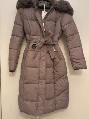 Lång brun dunjacka med fuskpäls köpt för 1200kr använd 3 gånger helt ny  - Snygg lång brun dunjacka med bälte i midjan och stor huva med mjuk fuskpäls. Jackan har quiltad design och dragkedja framtill. Perfekt för kalla vinterdagar och ger en riktigt trendig look.