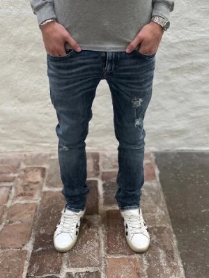 Mörkblå slitna slim fit jeans replay anbass - Säljer ett par mörkblå slim fit jeans med slitningar och hål på framsidan av benen. Jeansen har klassisk femficksdesign och snygg tvättad look. Jeansen är från replay och är i modellen anbass, riktigt snygga slitningar!