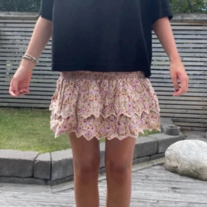 Blommig volangkjol med spetskant - Supersöt kort kjol med blommigt mönster i ljusa toner och volanger. Kjolen har en scallopad spetskant längst ner som ger en extra gullig touch. Perfekt för dig som gillar romantisk och feminin stil.