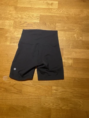 Svarta träningstights shorts Lululemon - Säljer ett par svarta träningstights shorts från Lululemon med hög midja och tight passform. Perfekta för gymmet eller löpning, tillverkade i ett stretchigt syntetmaterial som andas. Diskret logga på benet och bred linning för extra komfort.