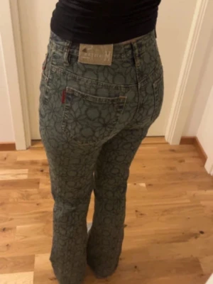 Blommiga Mönstrade bootcut jeans  - Säljer ett par unika blå jeans  med blommigt mönster över hela tyget. Modellen är bootcut med utsvängda ben. Jeansen har låg midja. De når precis ner till golvet på mig (166cm)