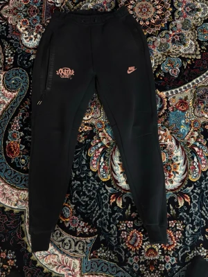 Nike syna world byxor - Säljer ett par svarta mjukisbyxor från Nike med rosa logga och grafiskt tryck på vänster ben. Byxorna har dragsko i midjan, ribbade muddar vid benslut och dragkedjeförsedd ficka. Perfekta för chill eller streetwear-stil.