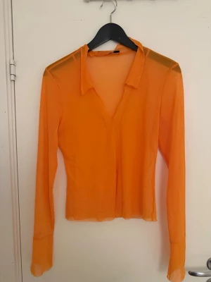 Transparent orange meshblus - Trendig orange meshblus med lång ärm och klassisk krage. Blusen är transparent och har en figurnära passform som ger en cool och modern look. Perfekt att styla lager på lager eller med ett snyggt linne under.