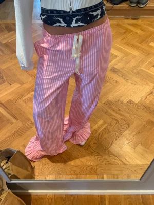 Rosa randiga pyjamasbyxor - Säljer ett par rosa pyjamasbyxor med vita vertikala ränder och breda ben. Byxorna har en bekväm resår i midjan och ett vitt knytband framtill. Perfekta för mysiga hemmakvällar eller lata morgnar.