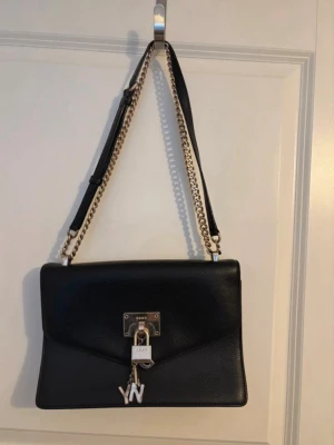Svart axelväska från DKNY - Snygg svart axelväska från DKNY i slätt läder med guldfärgad kedjerem och dekorativt hänglås framtill. Väskan har en stilren rektangulär form och detaljer i metall, inklusive DKNY-logga och berlock. Perfekt för dig som gillar trendiga accessoarer. Kolla min profil för fler plagg! 🥰