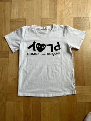 Vit Comme des Garçons PLAY t-shirt - Säljer en vit t-shirt från Comme des Garçons PLAY med det ikoniska svarta hjärtat och loggan tryckt på bröstet. Klassisk rund hals och korta ärmar. Tillverkad i mjuk bomull som känns skön mot huden. Perfekt för en clean och stilren look. Otroligt bra skick förutom en liten defekt på bokstaven C i loggan. Vid frågor eller funderingar är det bara att säga till Mvh Olof.