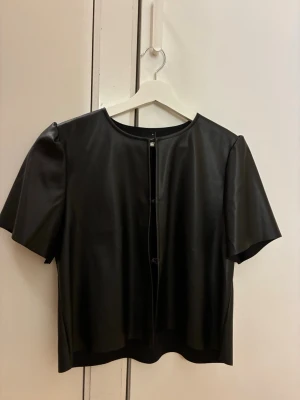 Svart PU-blus  - Trendig svart blus i PU-material med rund halsringning och korta, vida ärmar. Blusen har en rak passform och knäppning framtill. Använd 2ggr! Perfekt att ha till festen🌟