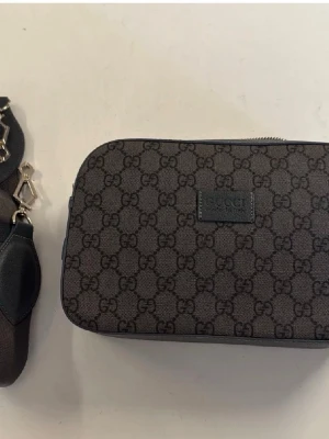 Gucci axelväska med GG-mönster - Snygg axelväska från Gucci i brunt tyg med klassiskt GG-monogram över hela väskan. Rektangulär form med svart läderdetalj och silverfärgade metalldelar. Märkeslogga på framsidan. Perfekt för dig som gillar exklusiva accessoarer.