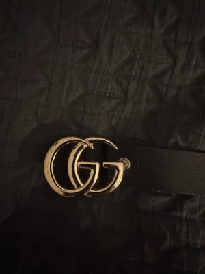 Svart skärp med GG-spänne i guld - Snyggt svart bälte från Gucci med ikoniskt GG-spänne i guld. Tillverkat i 100% äkta skinn och passar perfekt till jeans eller byxor för en trendig look. Bältet har en klassisk design som lyfter vilken outfit som helst.