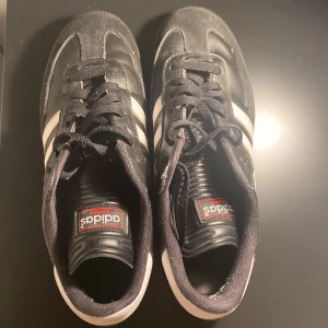 Adidas Samba svart/vit strl 42 - Klassiska Adidas Samba sneakers i svart skinn och mocka med vita ränder på sidorna och svart snörning. Snygg retrostil med låg profil och platt sula. Perfekta för dig som gillar streetwear och tidlös design.