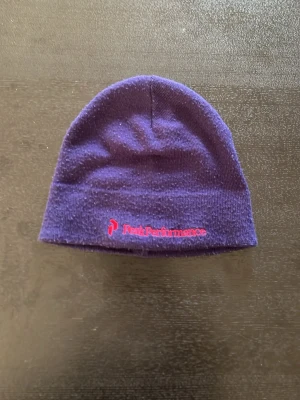 Lila mössa från Peak Performance - Snygg lila mössa från Peak Performance med broderad logga i rött framtill. Klassisk modell i mjukt stickat material som sitter skönt på huvudet. Perfekt för kalla dagar och enkel att matcha med olika outfits.