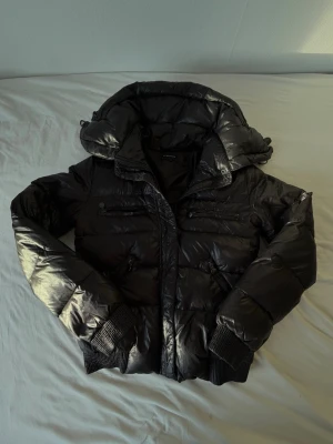 Svart pufferjacka från Reserved - Snygg svart pufferjacka från Reserved med glansig finish, och flera dragkedjefickor framtill. Jackan har en dold huva och är perfekt för kalla vinterdagar. 