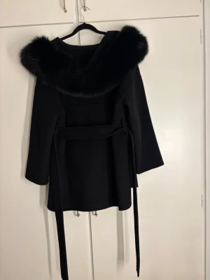 Svart kappa med äktapäls och knytskärp - Stilren svart kappa med stor huva kantad av äkta päls . Kappan har ett långt knytskärp i midjan och vida ärmar för en modern siluett. Perfekt för kalla dagar när du vill känna dig både varm och snygg.