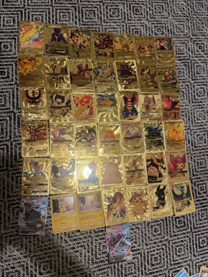 Pokémonkort samling guld & holo - Samling med Pokémonkort, mestadels guldfärgade samlarkort med motiv av olika Pokémon och tränare. Några kort är holografiska och färgglada. Korten är rektangulära och tillverkade av plast eller tjockt papper. Perfekt för dig som gillar Pokémon och vill ha unika kort i din samling.