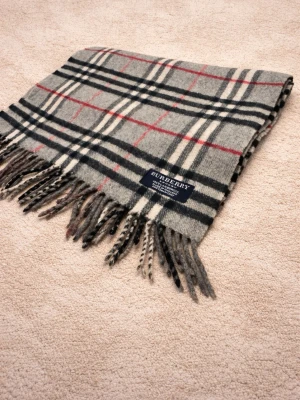 Grå rutig halsduk från Burberry - Klassisk halsduk från Burberry i grått med ikoniskt rutmönster i svart, vitt och rött. Tillverkad i mjuk ull och har fransar i båda ändar. Perfekt accessoar för att lyfta din outfit med en tidlös och lyxig känsla. 