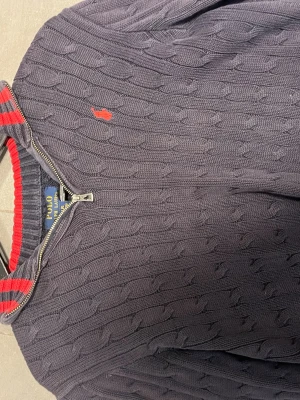 Mörkblå kabelstickad tröja Polo Ralph Lauren - Kabelstickad mörkblå tröja från Polo Ralph Lauren med dragkedja och hög krage. Tröjan har röda detaljer på kragen och en röd broderad logga på bröstet. Tillverkad i mjukt material, perfekt för kyliga dagar.