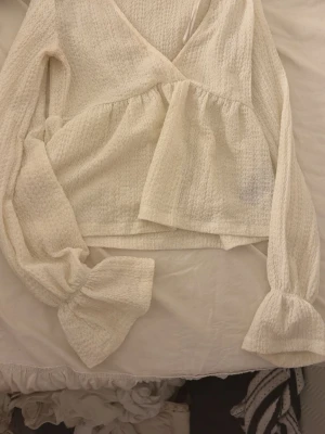 Vit omlottblus från Gina Tricot - Supersöt vit omlottblus från Gina Tricot i strukturerat tyg. Blusen har långa puffiga ärmar med resår vid ärmslut och en volang nedtill. Perfekt för att skapa en romantisk look och lätt att matcha med jeans eller kjol.