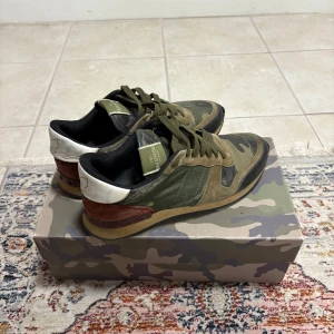 Valentino camo sneakers i mocka - Snygga sneakers från Valentino Garavani med kamouflagemönster i grönt, brunt och rött. Skorna har detaljer i mocka, läder och textil samt en vit hälkappa och röda partier vid hälen. Klassisk låg modell med gröna skosnören och gummisula. Det finns några defekter därav priset. Kvitto, box och skopåsar tillkommer.